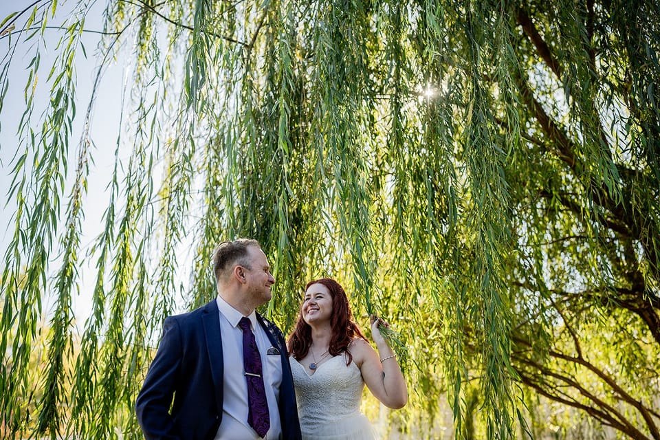Steve in a navy suit and Katie in a white dress smiling at each other under a weeping willow, sunlight through the leaves