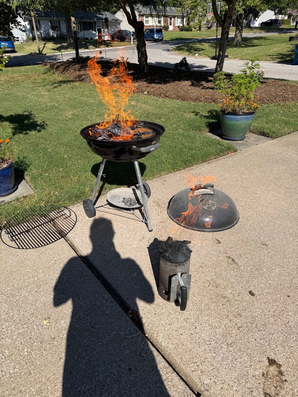 A charcoal grill on a driveway with flames rising off the coals, its lid and a chimney starter beside it
