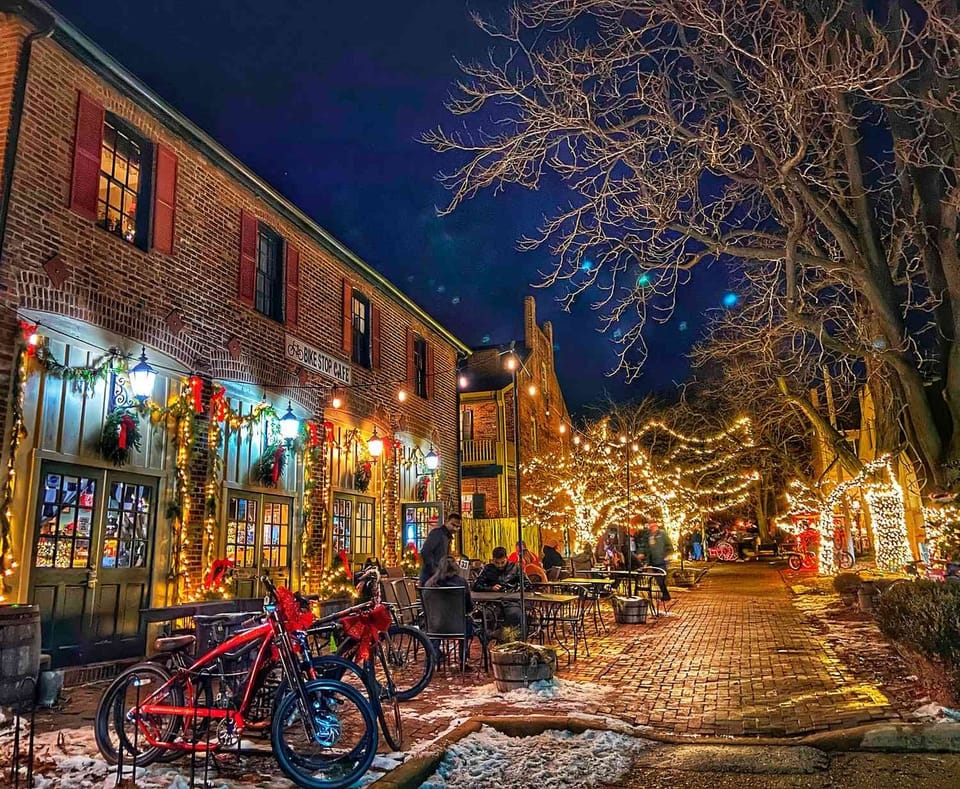Main Street in St. Charles at night at Christmas: a brick building hung with wreaths and garlands, bicycles with red bows, and trees wrapped in white lights along a brick walk