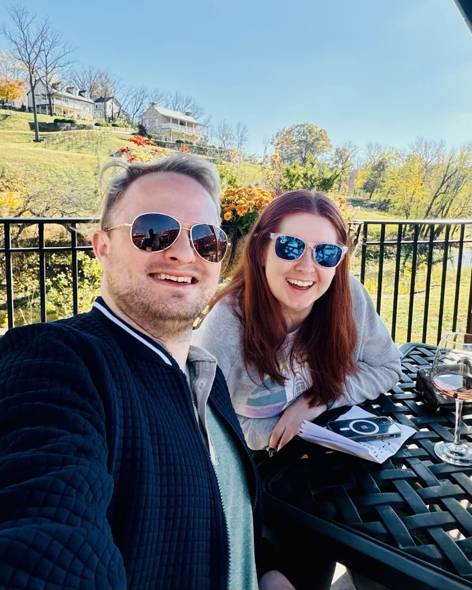 Steve and Katie smiling in sunglasses at a table on a vineyard terrace, autumn trees and a hillside behind them